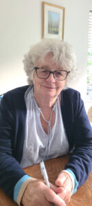 An elderly woman with short, curly white hair and black-rimmed glasses sits at a wooden table, holding a pen in her hands. She is wearing a white blouse with delicate embroidery and a navy blue cardigan with light blue cuffs. She has a warm, gentle smile and is looking directly at the camera. Behind her, a framed landscape painting hangs on a white wall, and a window with blinds allows natural light to brighten the room.